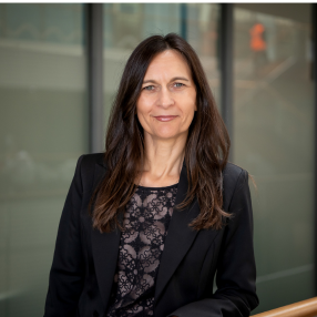 Professional headshot of a woman in a black blazer, photographed in a modern office building.