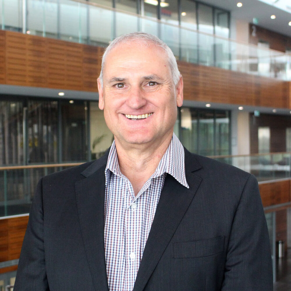 Professional headshot of a smiling man in a dark blazer and checked shirt, photographed in a modern office building.