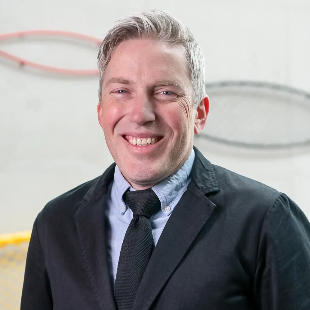 Professional headshot of a smiling man in a dark blazer and tie, photographed in front of a light-coloured wall with circular artwork.
