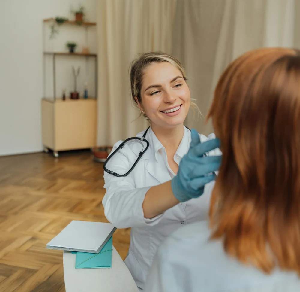 A smiling female doctor wearing a white coat, stethoscope and gloves speaking with a patient during a consultation.