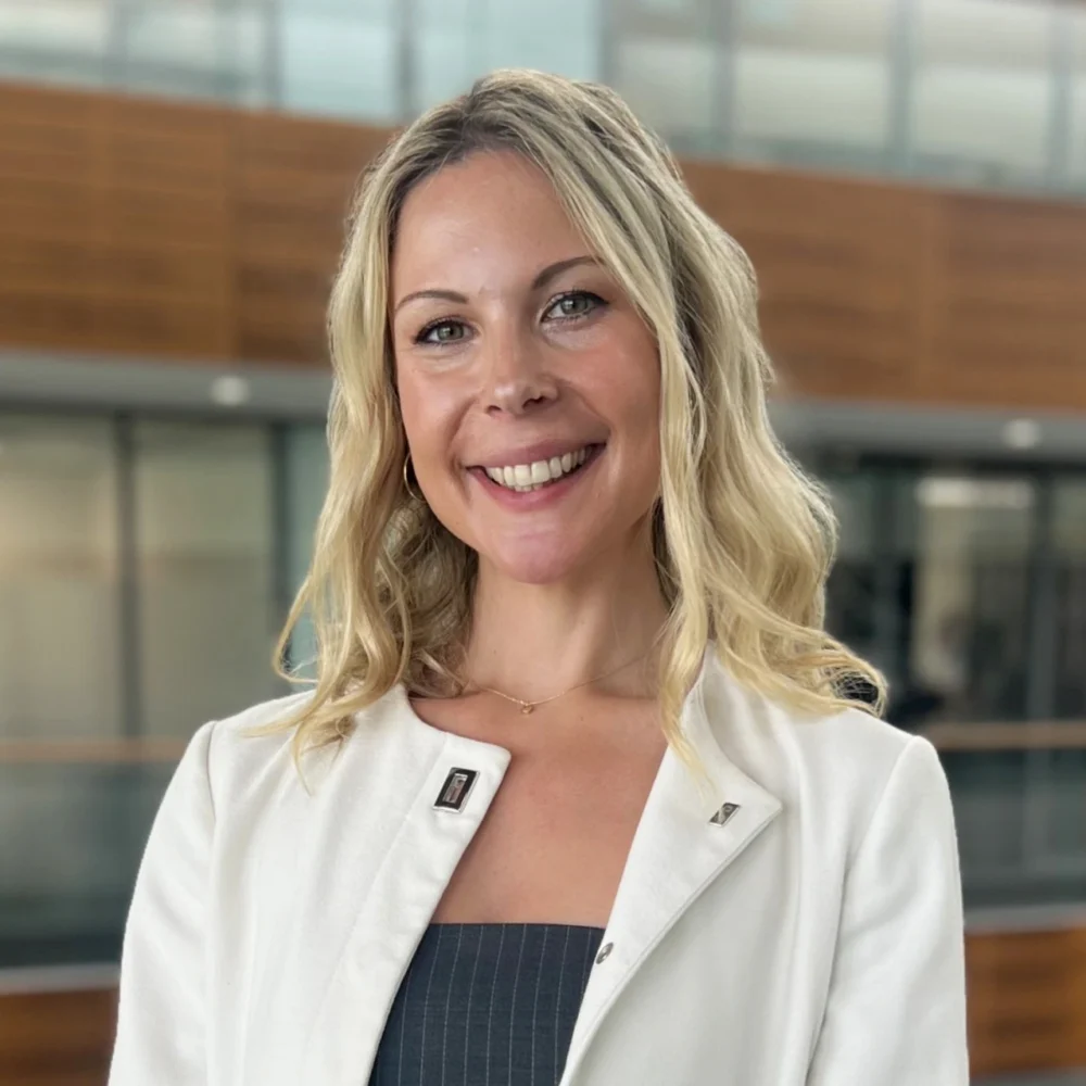 Professional headshot of a smiling woman in a white blazer, photographed in a modern office building.