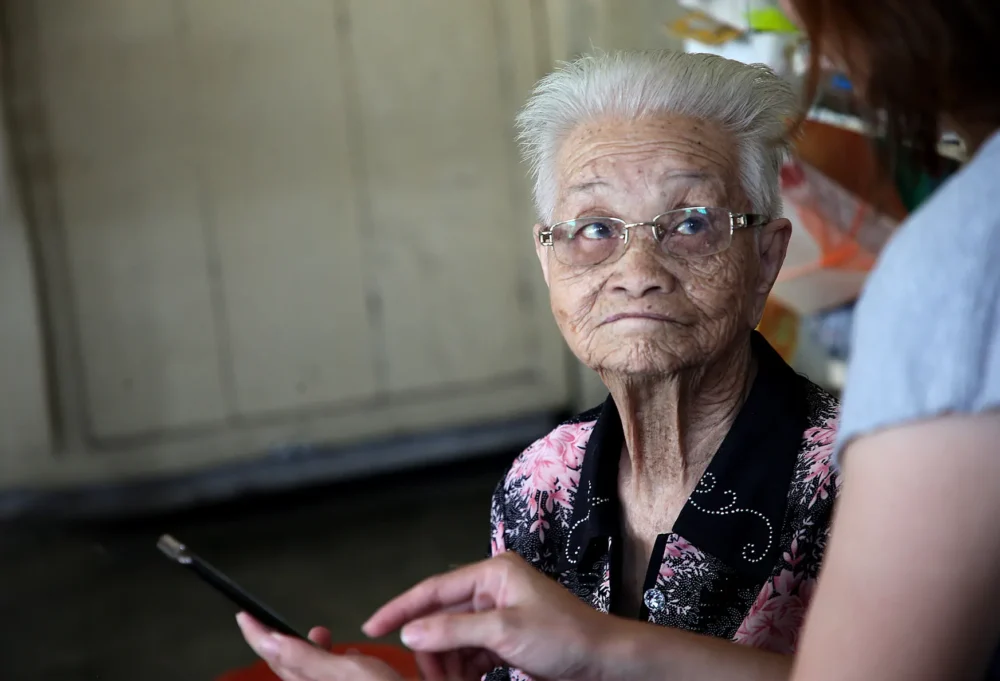 An elderly woman wearing glasses looking attentively at another person holding a tablet or phone during what appears to be a care or support visit.